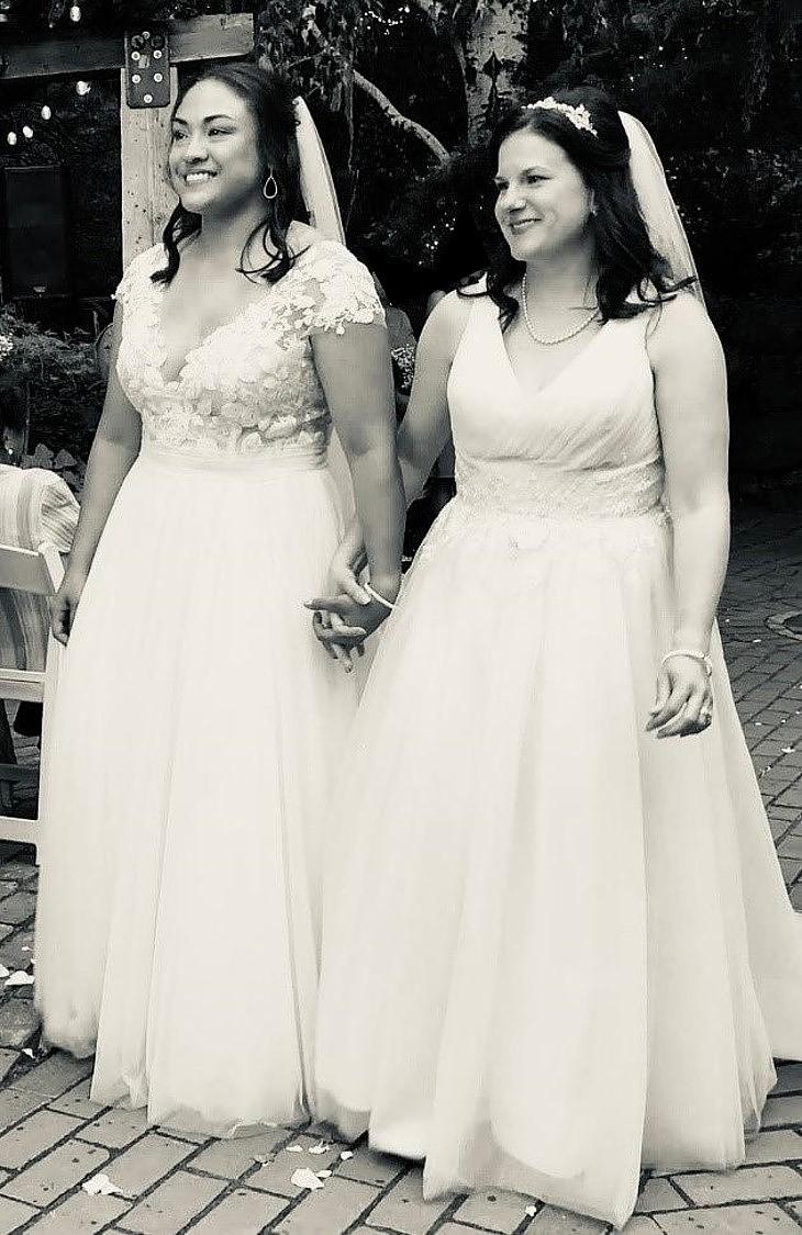 Kristin and her wife are dressed in wedding gowns, holding hands and smiling. 